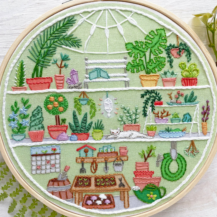 A circular embroidery hoop containing a colorful cross-stitch design of a greenhouse filled with potted plants, shelves, gardening tools, and small decorative elements, displayed on a white wood surface with greenery props.