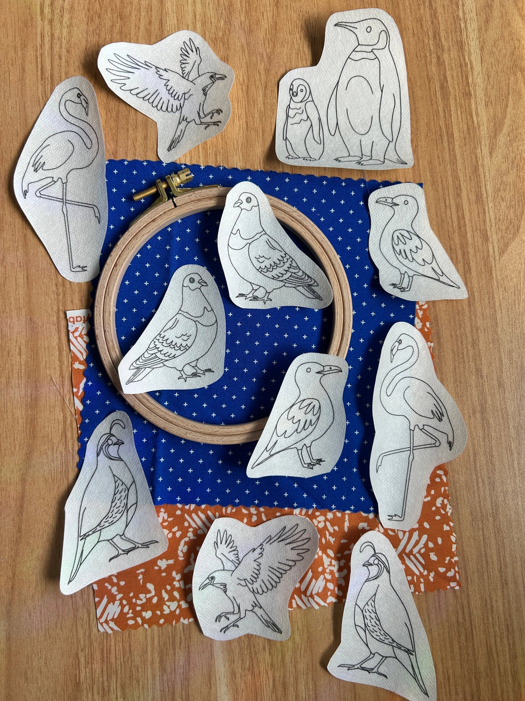Odd Birds Stick & Stitch Embroidery Patterns featuring various bird designs outlined on fabric pieces arranged around an embroidery hoop atop patterned blue and orange fabric, ideal for modern needlework and embroidery thread projects.