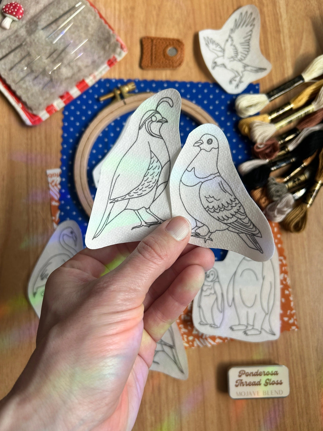 Hand holding bird pattern templates over a blue embroidery hoop surrounded by embroidery threads, needles, and sewing supplies for Odd Birds Stick & Stitch Embroidery Patterns.