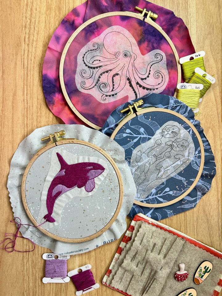 PNW Animals Stick & Stitch Embroidery Patterns displayed in hoops featuring an octopus, orca whale, and otters with vibrant thread and needlework supplies on wooden surface.
