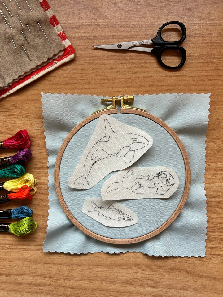 PNW Animals Stick & Stitch Embroidery Patterns featuring outlined designs of an orca, otter, and fish on fabric with embroidery floss, scissors, and needles for modern needlework craft projects.