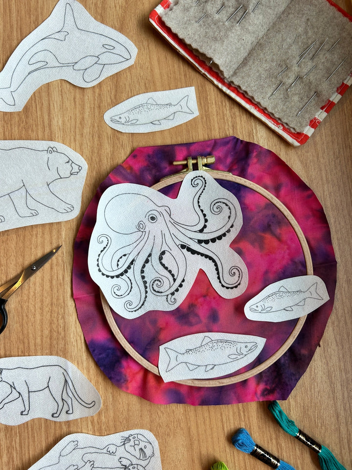 PNW Animals Stick & Stitch Embroidery Patterns featuring outlined animal designs on fabric, with an embroidery hoop, needles, scissors, and colorful threads arranged on a wood surface.