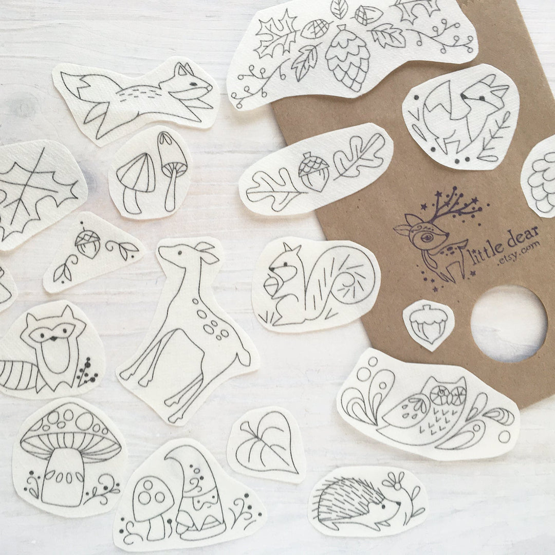 A collection of cute, hand-drawn forest-themed stickers with black line art on white backing, arranged on a light wood surface next to a brown kraft paper card printed with the brand name 'Little dear'.